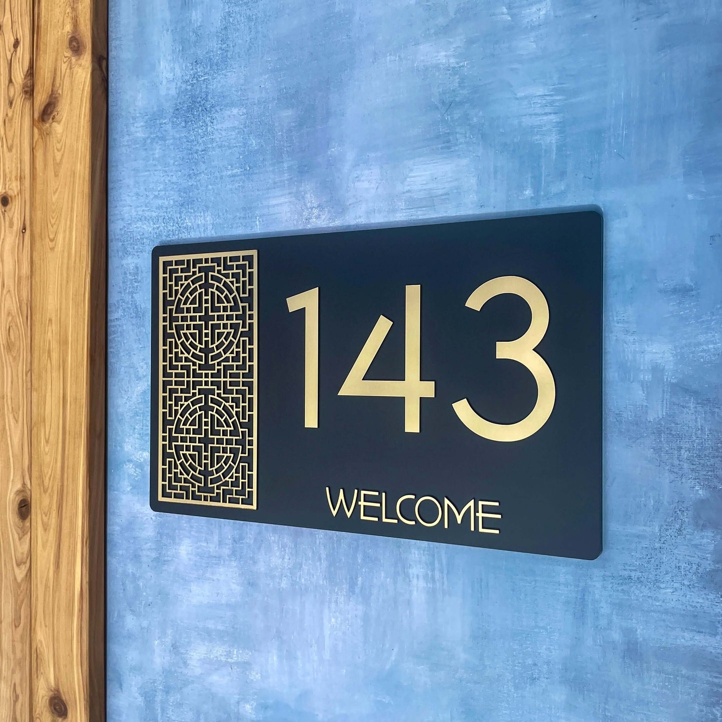 Decorative house number sign '143' in a modern design on a blue wall, with intricate patterns and 'Welcome' text below.