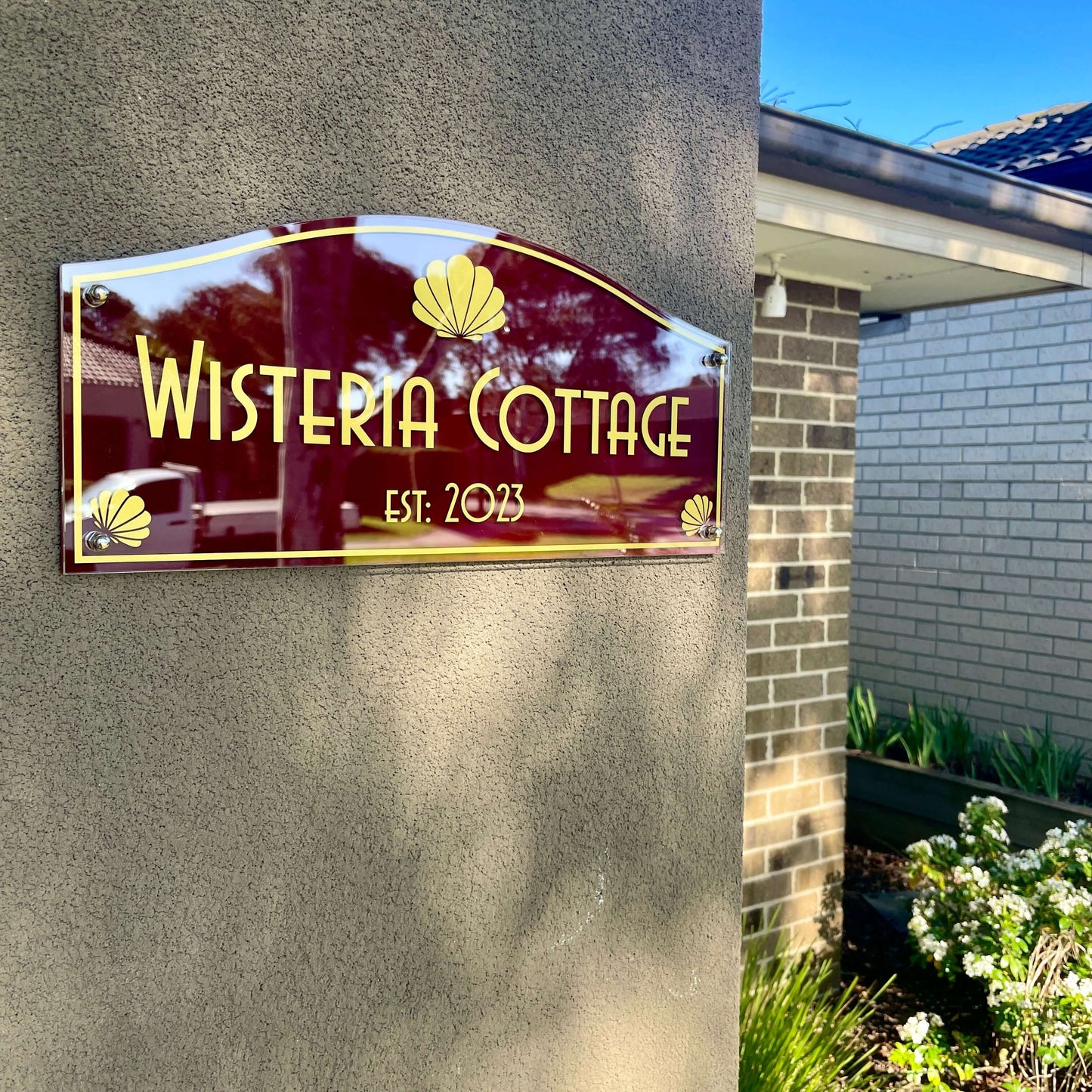 Gold and burgundy house sign reading 'Wisteria Cottage Est. 2023' mounted on a wall.