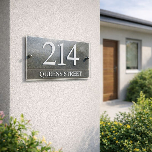 Urban 5 House number sign '214 Queens Street' in graphite and white, landscape style, on a house exterior with a door and garden in the background.