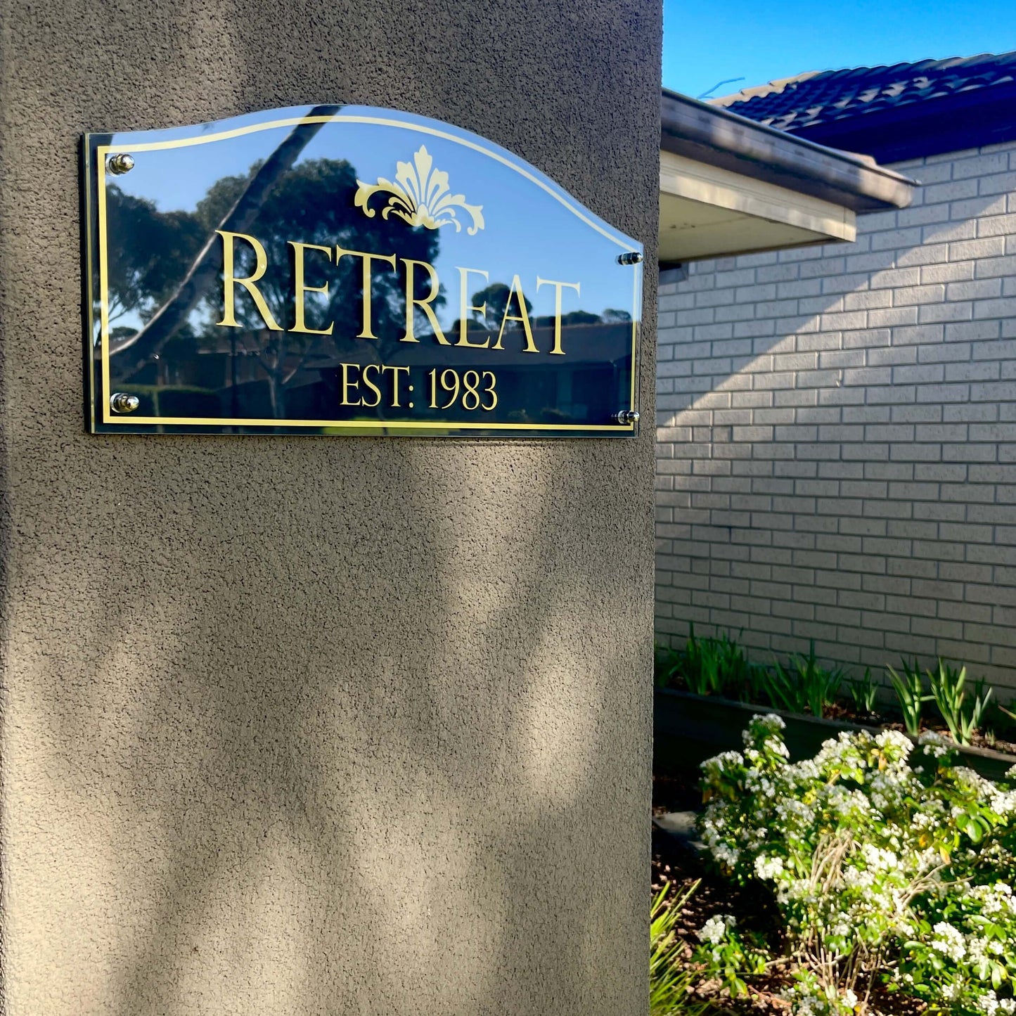 Clear outdoor sign with 'RETREAT EST: 1983' mounted on a textured wall beside a garden, reflecting blue sky and trees.