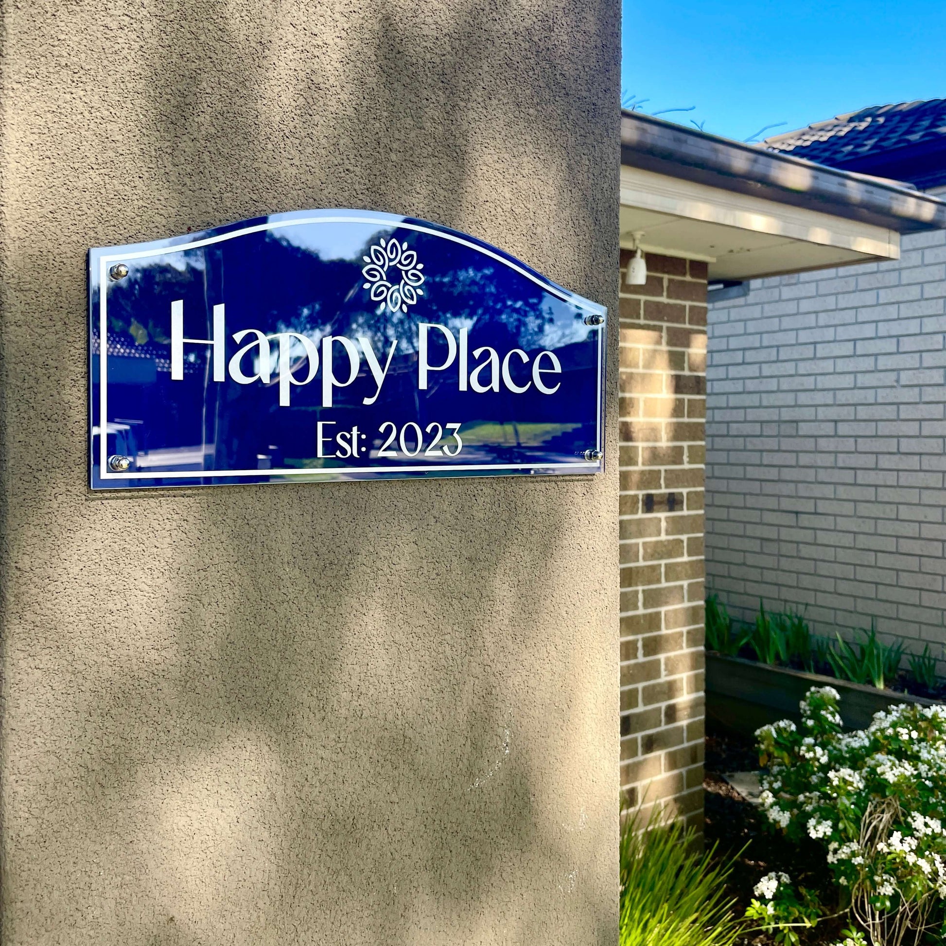 House with blue 'Happy Place' sign, est. 2023, against brick wall in garden setting.