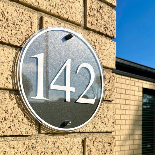 Contemporary SW2 house number sign in round shape, mounted on brick wall under blue sky, featuring number 142.
