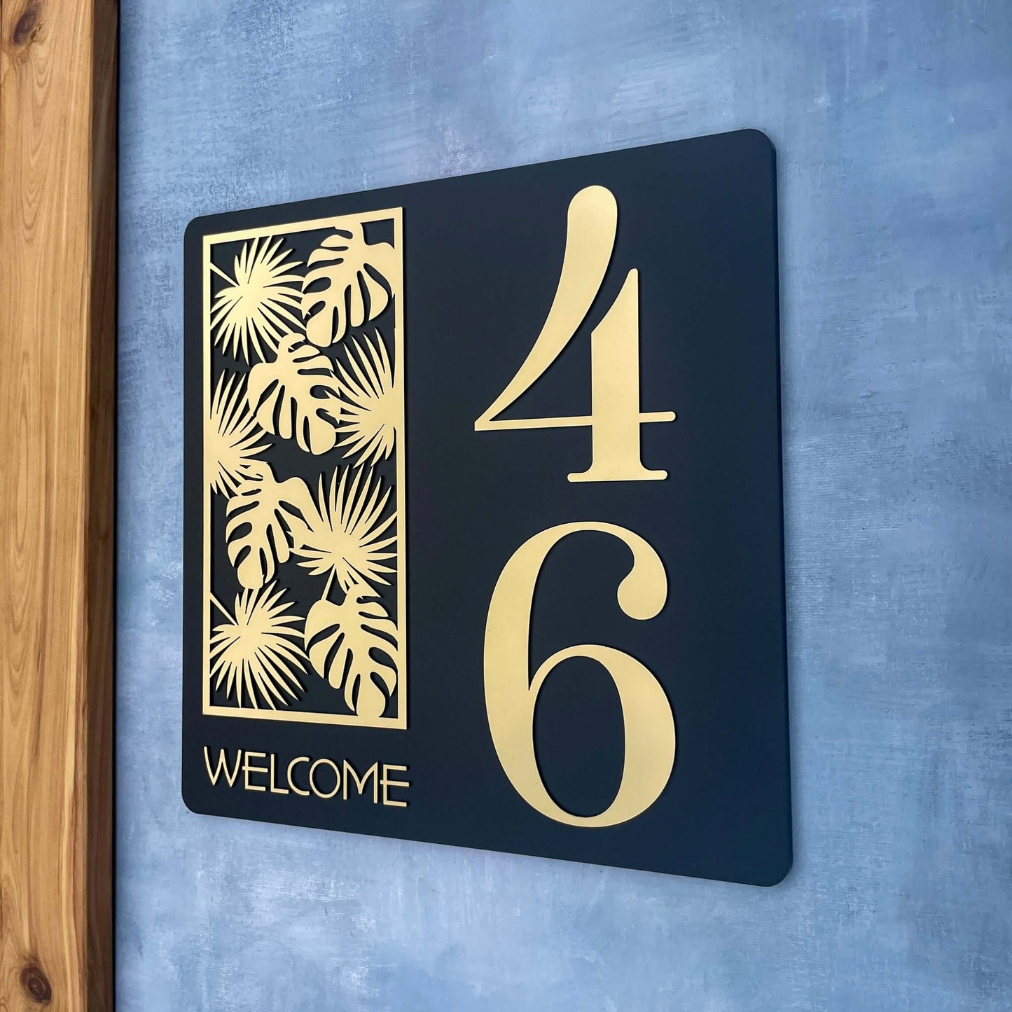 Stylish house number sign with gold tropical design and large numbers 46, mounted on a textured wall.